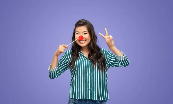 red nose day, party props and photo booth concept concept - happy woman with clown nose showing peace hand sign over violet background