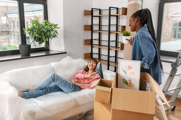 moving, people and real estate concept - happy smiling woman unpacking box and her friend resting on sofa at new home