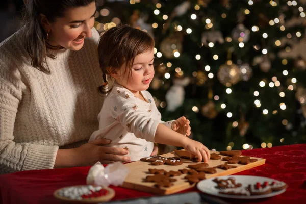 family, cooking and winter holidays concept - happy mother and baby daughter with icing in baking bag decorating gingerbread cookies at home on christmas