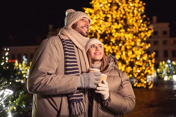 winter holidays, hot drinks and people concept - happy young couple with takeaway coffee cups over christmas tree lights in evening city
