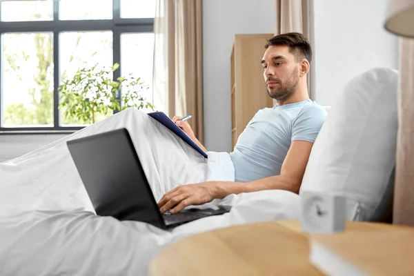 people, technology and remote job concept - man with laptop computer and clipboard working in bed at home bedroom