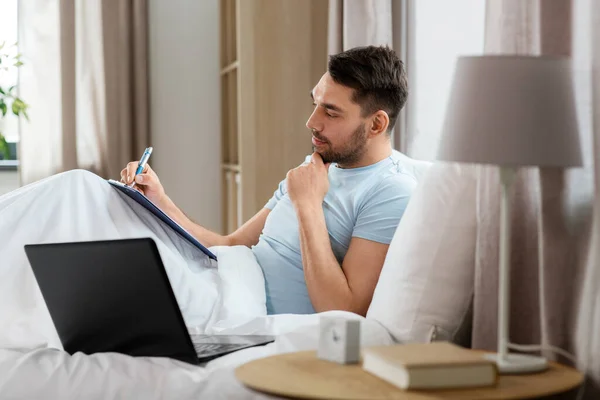 リモートジョブの概念 ラップトップコンピュータとクリップボードを持つ男は 自宅の寝室でベッドで働いています — ストック写真