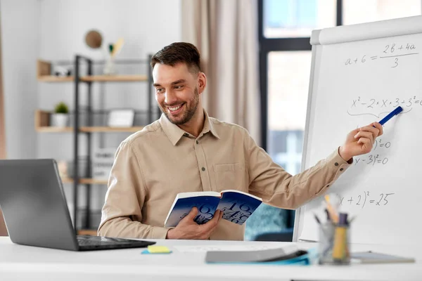 distant education, school and remote job concept - happy smiling male math teacher with laptop compute and book showing equation on flip chart having online class or video call at home office