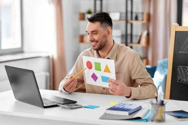 distance education, primary school and remote job concept - happy smiling male teacher with laptop and picture of geometric shapes in different colors having online class at home office