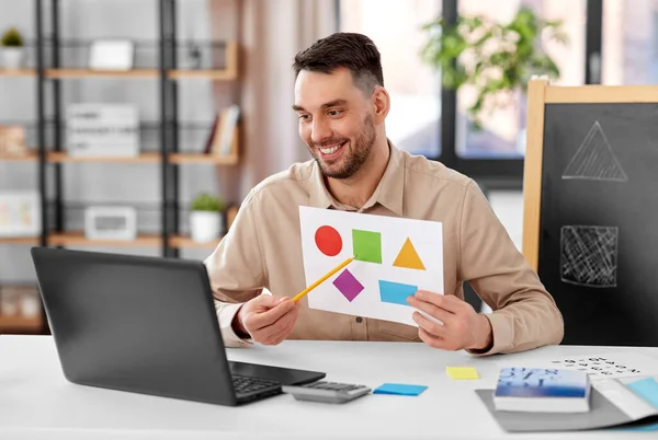 distance education, primary school and remote job concept - happy smiling male teacher with laptop and picture of geometric shapes in different colors having online class at home office