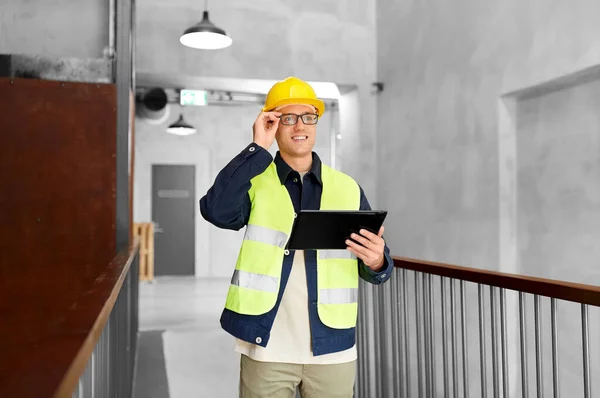 architecture, construction business and building concept - happy smiling male supervisor in helmet and safety west with tablet pc computer at office