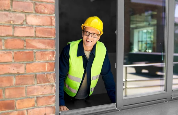 construction business and building concept - happy smiling male builder in helmet and safety west looking out window