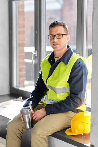 architecture, construction business and people concept - happy smiling male builder or architect with tumbler or thermo cup sitting on window sill at office