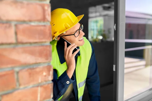 construction business and building concept - smiling male builder in helmet and safety west calling on smartphone and looking out window
