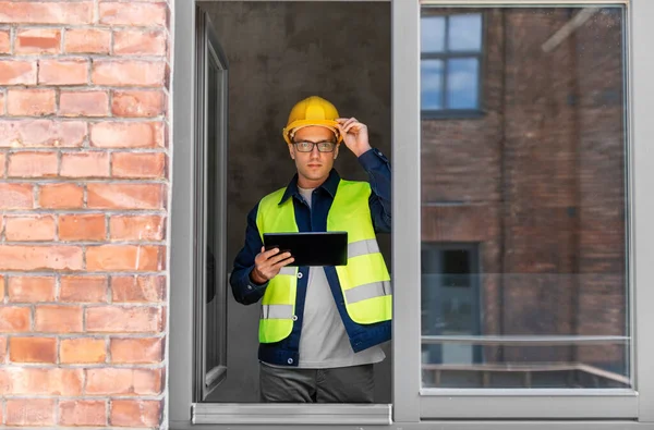 architecture, construction business and building concept - male architect in helmet and safety west with tablet pc computer in window