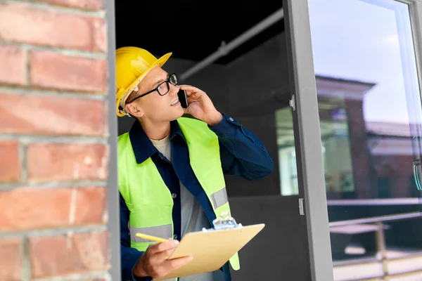 construction business and building concept - happy smiling male builder in helmet and safety west with clipboard calling on smartphone at window