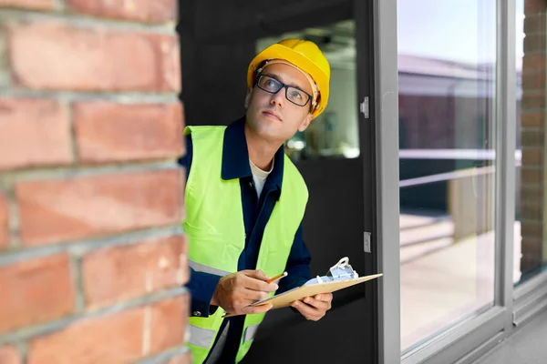 construction business and building concept - male builder in helmet and safety west with clipboard and pencil looking out window