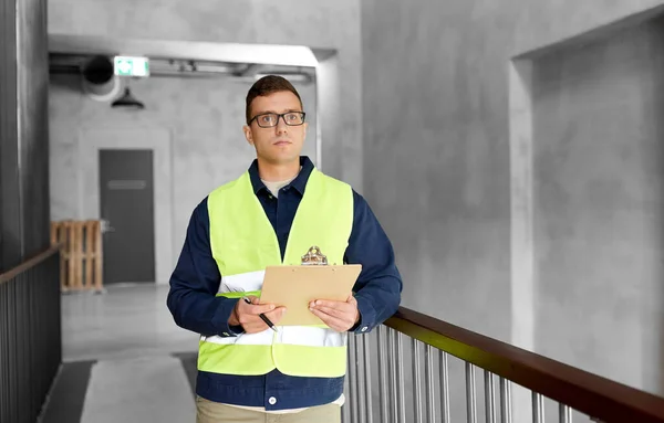 architecture, construction business and building concept - male supervisor in safety west with clipboard working at office