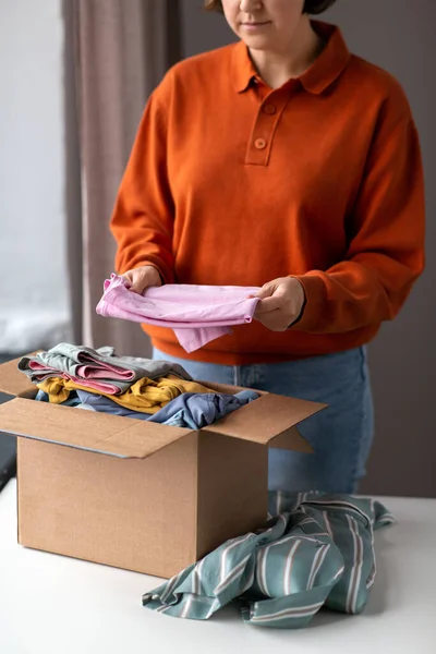 charity, donation and volunteering concept - young woman puts clothes in cardboard box at home