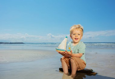Küçük çocuk deniz kıyısında oturan oyuncak yelkenliyle oynuyor.