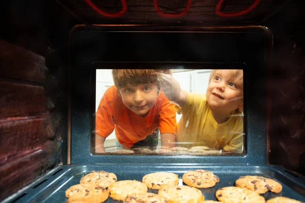 小さな子供たち2人の兄弟はオーブンのドアガラスを見てクッキーが中で焼かれている — ストック写真