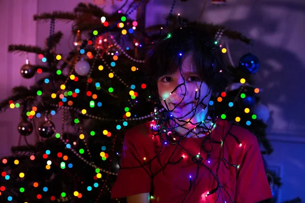 Chłopiec Oświetlonymi Światłami Girlandy Nad Twarzą Pobliżu Choinki — Zdjęcie stockowe