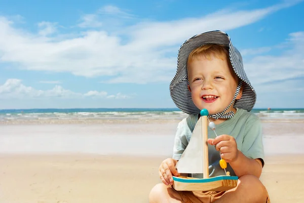 度假时 小男孩戴着巴拿马式的帽子 坐在海滩上玩抓帆游艇 — 图库照片