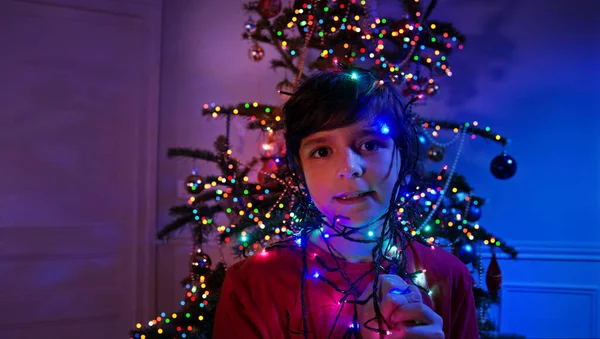 男孩站在装饰过的圣诞树旁 手上拿着装饰过的花环灯 — 图库照片