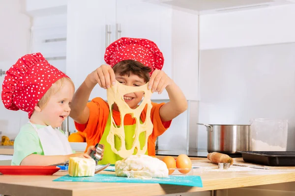 幸せな子供たちプレイ保持生地とともに穴切りクッキーを身に着けていますシェフ赤い帽子自宅キッチンに座って — ストック写真