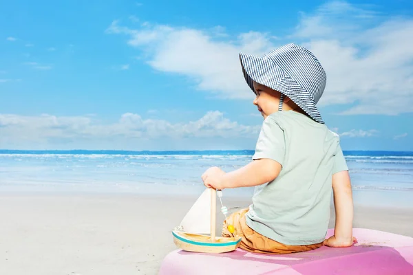 小さな男の子は後ろから海の景色と遊んでビーチの休暇のスーツケースに座って帆ボートヨットを保持再生します — ストック写真