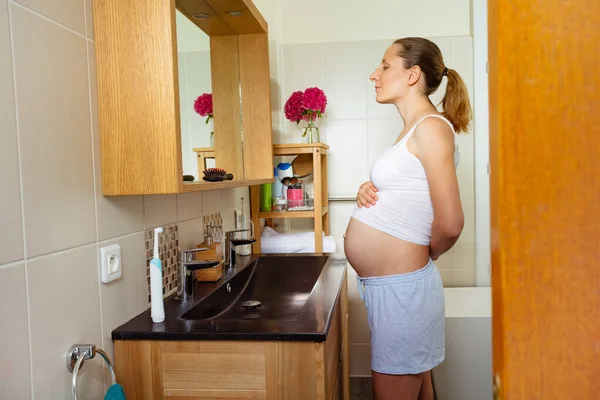 若いです妊娠中の女性立っていますバスルーム近くミラーとシンク見彼女の腹 — ストック写真