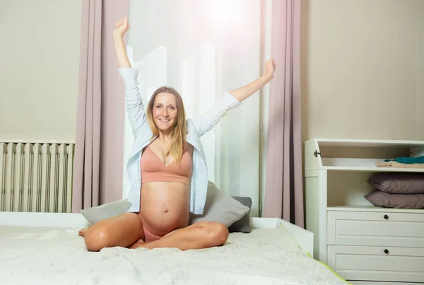 幸せなリラックス妊娠中の女性で白いシャツ座っています上のベッドストレッチ手と笑顔 — ストック写真