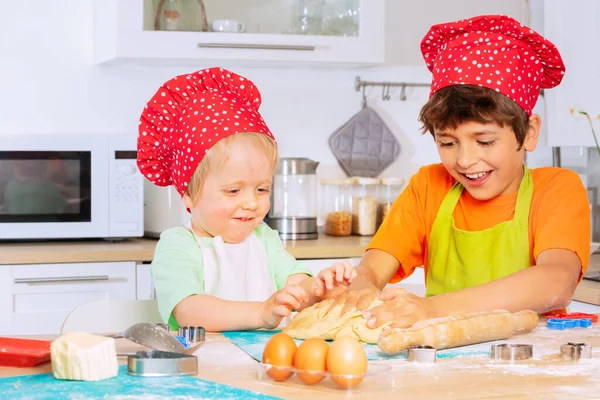 两个男孩手拿着厨师的红帽子 笑着坐在厨房桌旁 把面团揉碎了 — 图库照片