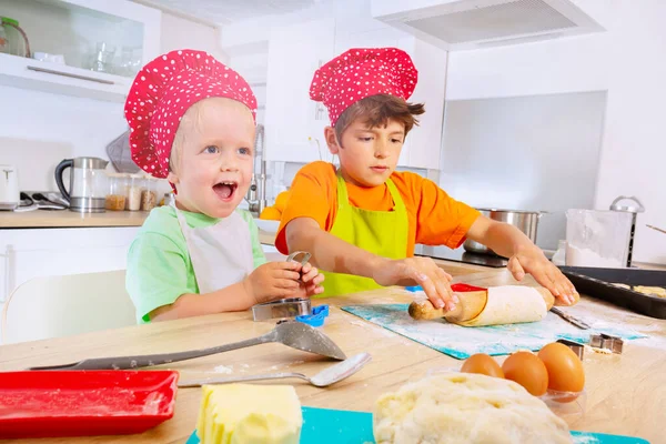 2幸せな小さな子供ロールアウト生地と準備クッキー上のキッチン身に着けているシェフの帽子 — ストック写真