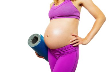 Spor kıyafetleri içinde koca göbekli hamile bir kadının yakın plan fotoğrafı. Elinde yoga arkadaşı, beyaz tenli, sırıtıyor.