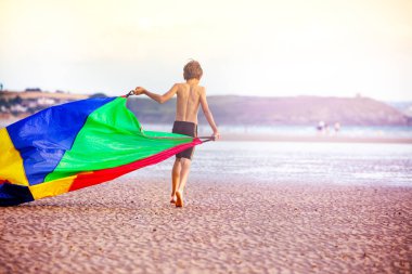 Genç çocuk deniz ve sahilde renkli bir paraşüt kumaşıyla yürüyor, arkadan manzaralı.