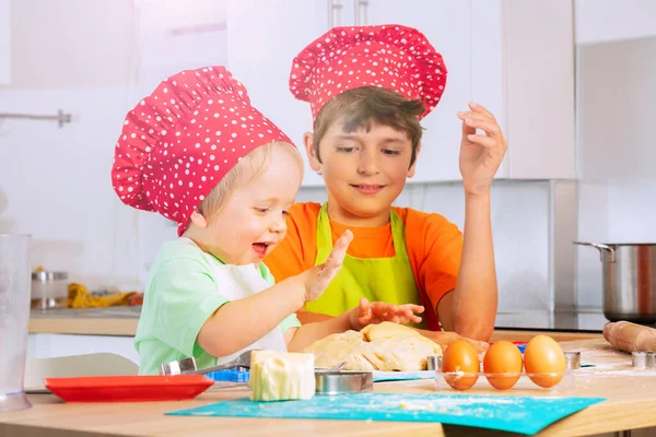 2人の子供がシェフの赤い帽子をかぶって手を使って生地を粉砕し キッチンのテーブルのそばに笑顔で座って — ストック写真