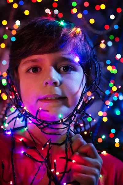 一个可爱的笑着的男孩的近照 他的花环灯火通明 在装饰过的圣诞树旁露出淡淡的微笑 — 图库照片