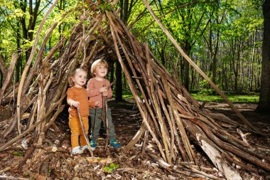 Two little kids, boy and a girl with pile of wood play in hut of branches or wickiup in the forest, summer activity at camp