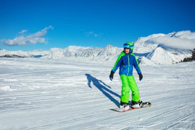 Çocuk, arka planda dağ sırasının zirvesiyle snowboard 'da yokuş aşağı gidiyor.