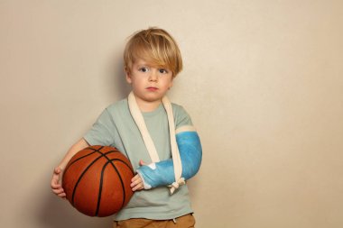 Üzgün, ağlayan, eli kırık, basketbol topu tutan küçük sarışın çocuk.