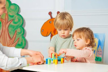 Mother and two little small children build tower with color blocks on the table clipart