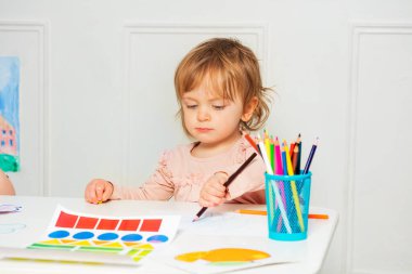 Little beautiful girl sit by the table and draw picture using pencil sitting with concentrated expression by the desk