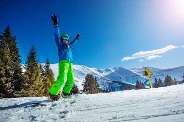 Snowboard 'daki çocuk, elleri havada, kış sporu kıyafeti, miğfer ve maskeyle kar dağlarının zirvelerinde duruyor.