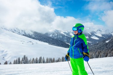Güneşli bir kış gününde, spor kıyafetiyle kayak pistinde durup dağları izleyen bir çocuğun yakın portresi.