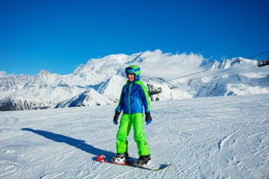Snowboard sporcusu gibi giyinmiş, kasklı ve maskeli arka planda Alp zirveleri olan bir çocuk.