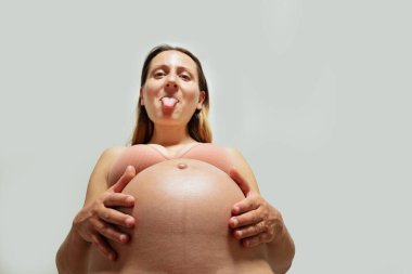 Funny playful pregnant woman stick out tongue look down holding belly