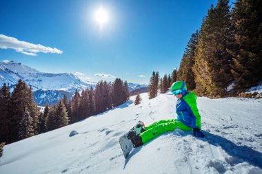 Snowboard 'lu çocuk kayak pistinde oturuyor. Dağların tepesinde, arka planda orman ve güneş.
