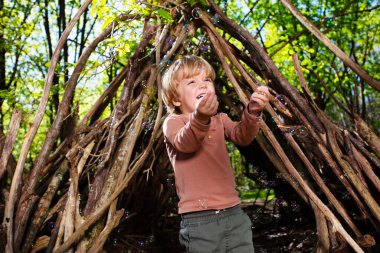 Happy little blond boy play with soap bubbles in hut of branches in the forest