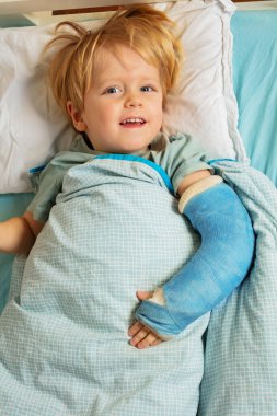 View from above of a little blond boy in the hospital bed with plaster cast on the broken hand, yet smile on the face