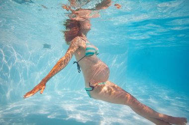 Açık havuzda su altında göbek egzersizi yapan hamile bir kadın.