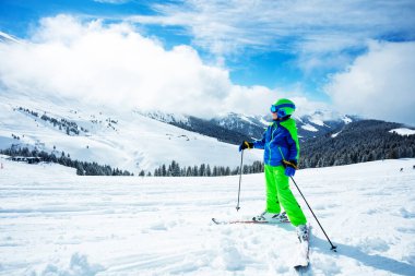 Little preteen boy stand on the ski track in sport outfit observing the mountain on sunny day at winter vacation