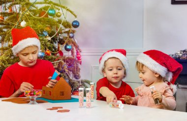 Bir grup çocuk Noel ağacının yanında Noel Baba şapkalarında oturan zencefilli kurabiye evini dekore ediyor.