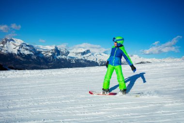 Snowboard 'daki çocuğun profil aksiyon fotoğrafı arka planda Alp dağlarının zirveleriyle yamaçtan aşağı kayıyor.