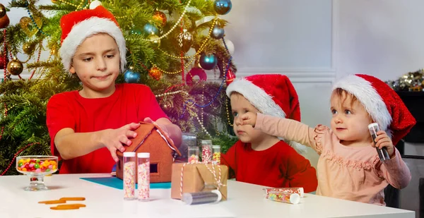 Kindergruppe Schmückt Lebkuchenhaus Mit Nikolausmützen Neben Weihnachtsbaum — Stockfoto